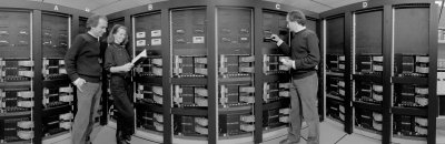 Standing in front of the outdated ACPMAPS parallel computer, Paul Mackenzie (left) talks to physicist Aida El-Khadra  as Mackenzie (again!!) inserts a storage tape. A panoramic photo scan made this scene possible in 1991.